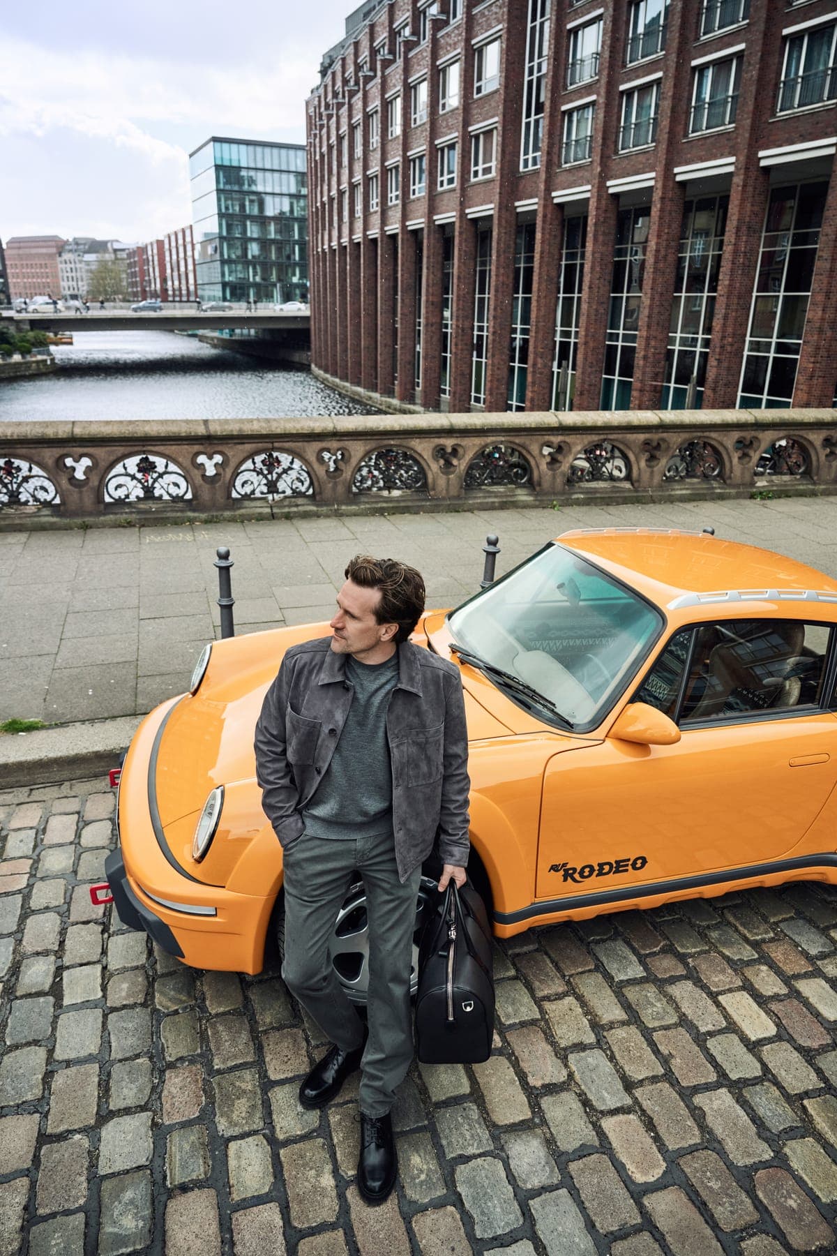 Man in NEFLYX denim leunend tegen een oranje sportwagen bij de haven